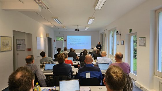 Ein Seminarraum mit mehreren Personen, die an einem KI-Workshop teilnehmen. Die Teilnehmer sitzen an Tischen mit Laptops und Notizblöcken, während ein Vortragender vorne steht und eine Präsentation auf einer Leinwand hält. Der Raum ist hell beleuchtet und verfügt über große Fenster, die einen Blick ins Grüne bieten.