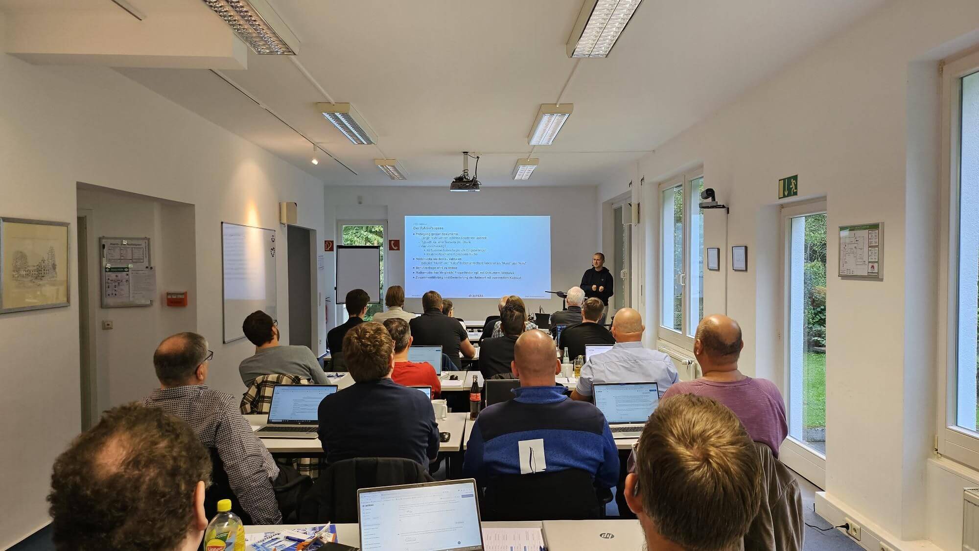 Ein Seminarraum mit mehreren Personen, die an einem KI-Workshop teilnehmen. Die Teilnehmer sitzen an Tischen mit Laptops und Notizblöcken, während ein Vortragender vorne steht und eine Präsentation auf einer Leinwand hält. Der Raum ist hell beleuchtet und verfügt über große Fenster, die einen Blick ins Grüne bieten.